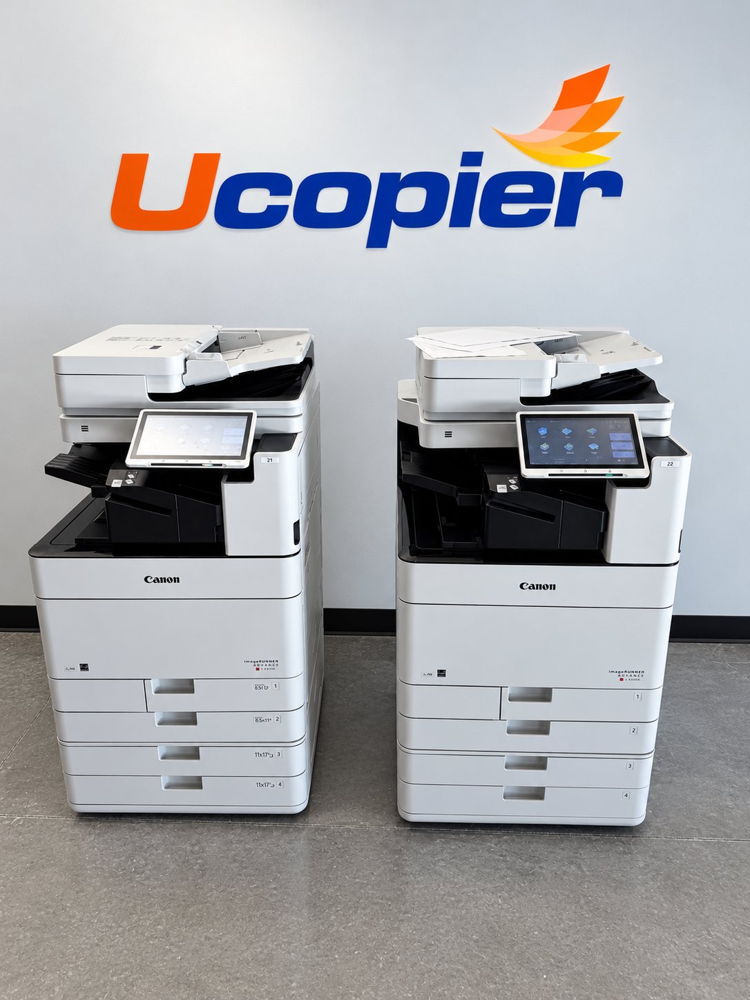 Two Canon multifunction copiers standing in front of the Ucopier logo on a white wall